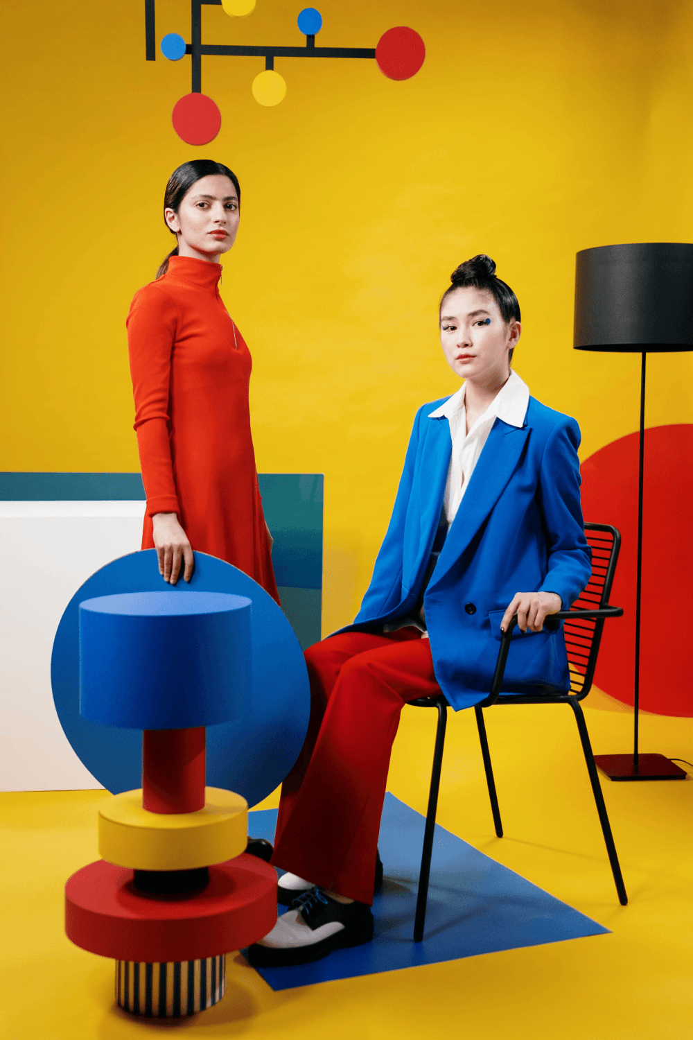 Twee personen die in de camera kijken. Een zit op een stoel de ander staat. Er zijn verschillende abstracte vormen te zien in de primaire kleuren blauw, geel en rood.