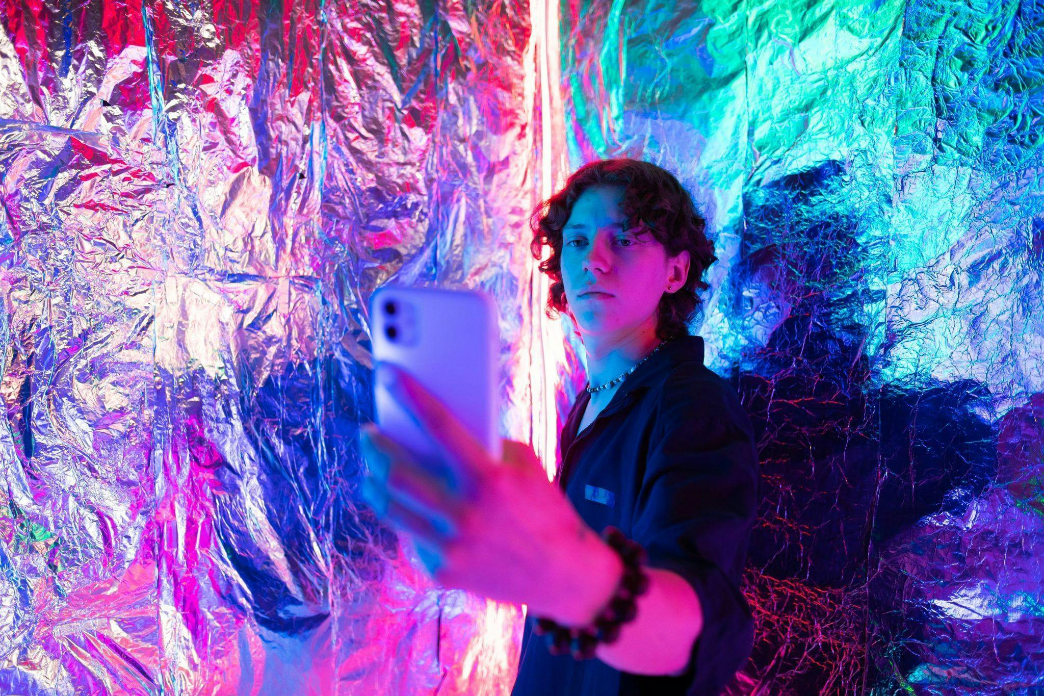 Jongere met donkere krullen staat met diens smartphone in de hand tegen een muur van glimmende folie. De jongere lijkt een selfie te maken.