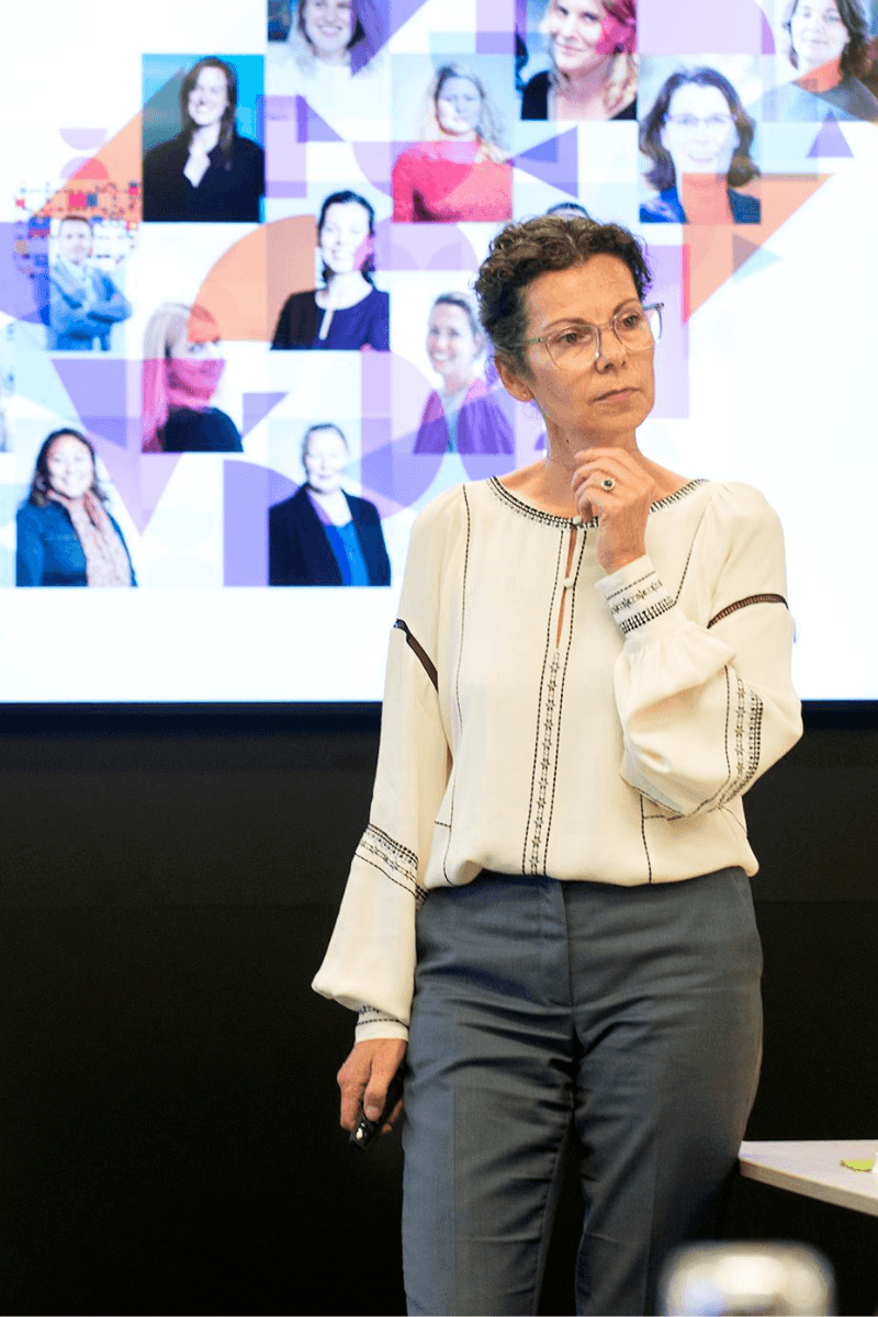 Maaike Verberk staat voor een presentatiescherm waarop het volledige DEN team te zien is. Ze draagt een grijze broek en lichte blouse. 