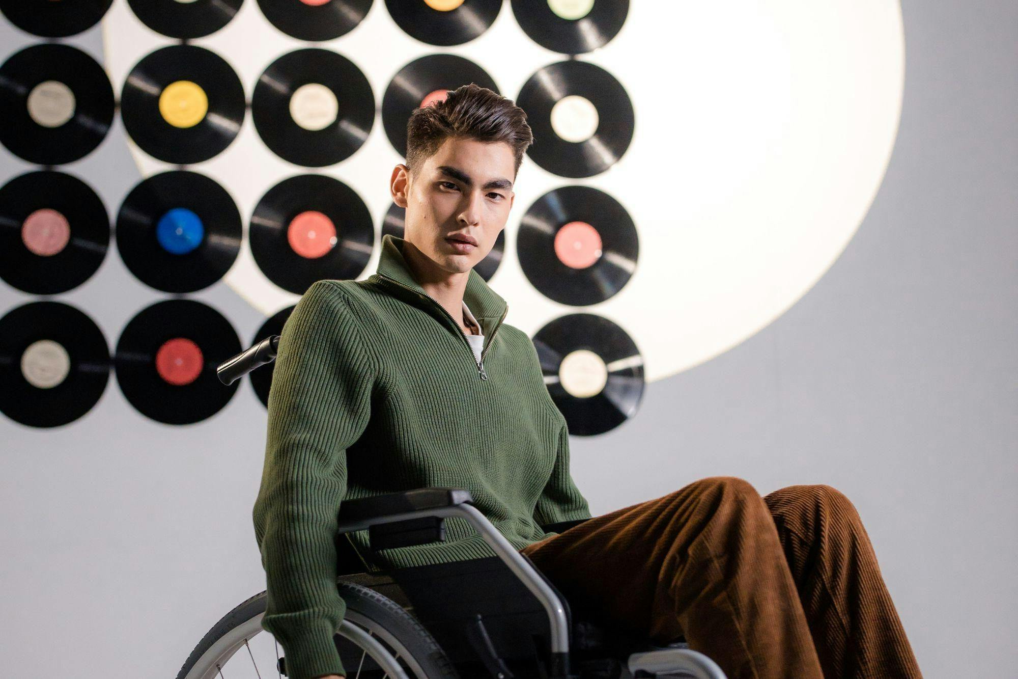 Persoon in een groene trui en bruine broek zit in zijn rolstoel voor een wand vol vinylplaten.