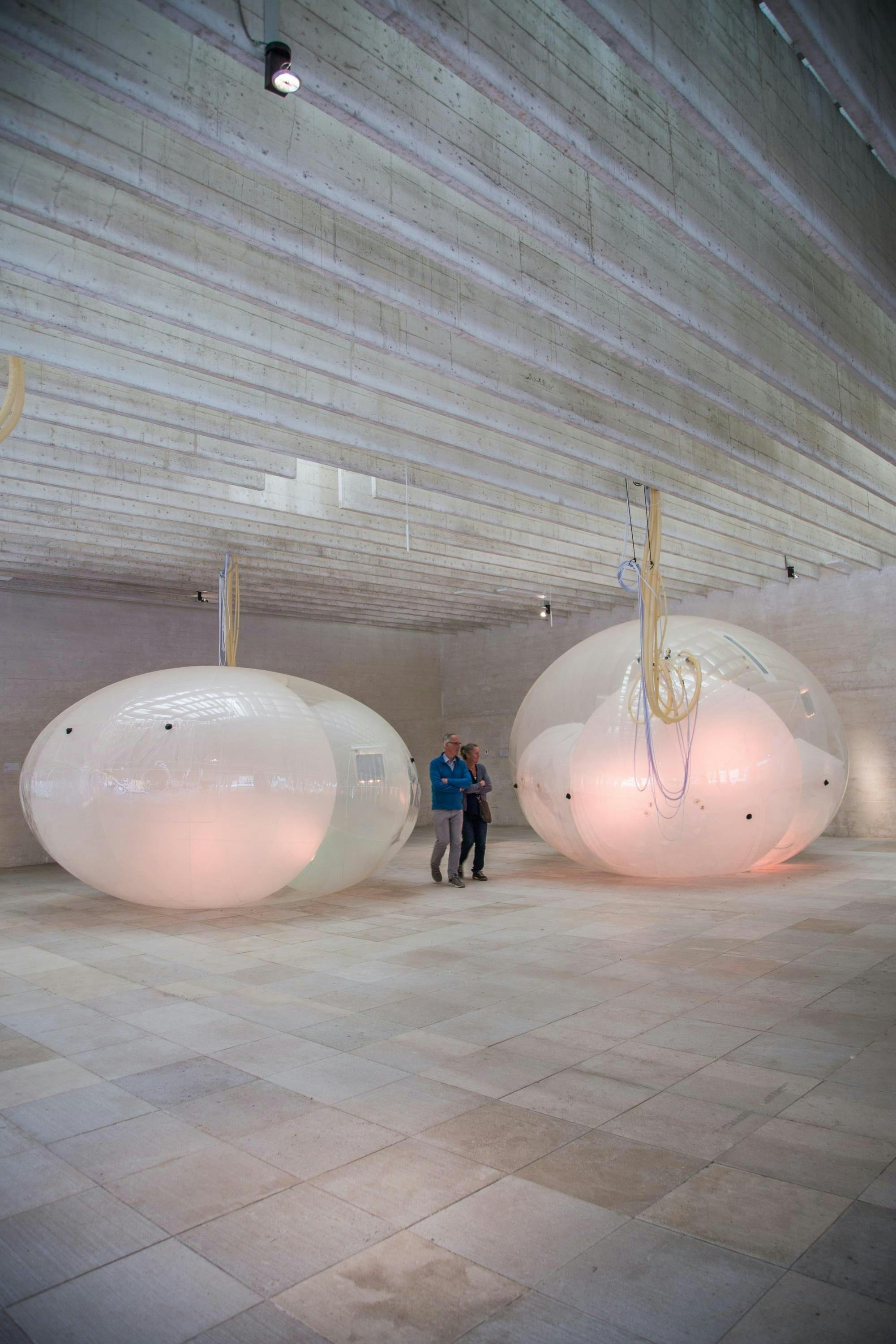 Twee mensen bekijken kunstobjecten. Het zijn een soort doorschijnende ballen met binnenin nog een paar ballen. De objecten zijn bevestigd aan het plafond.