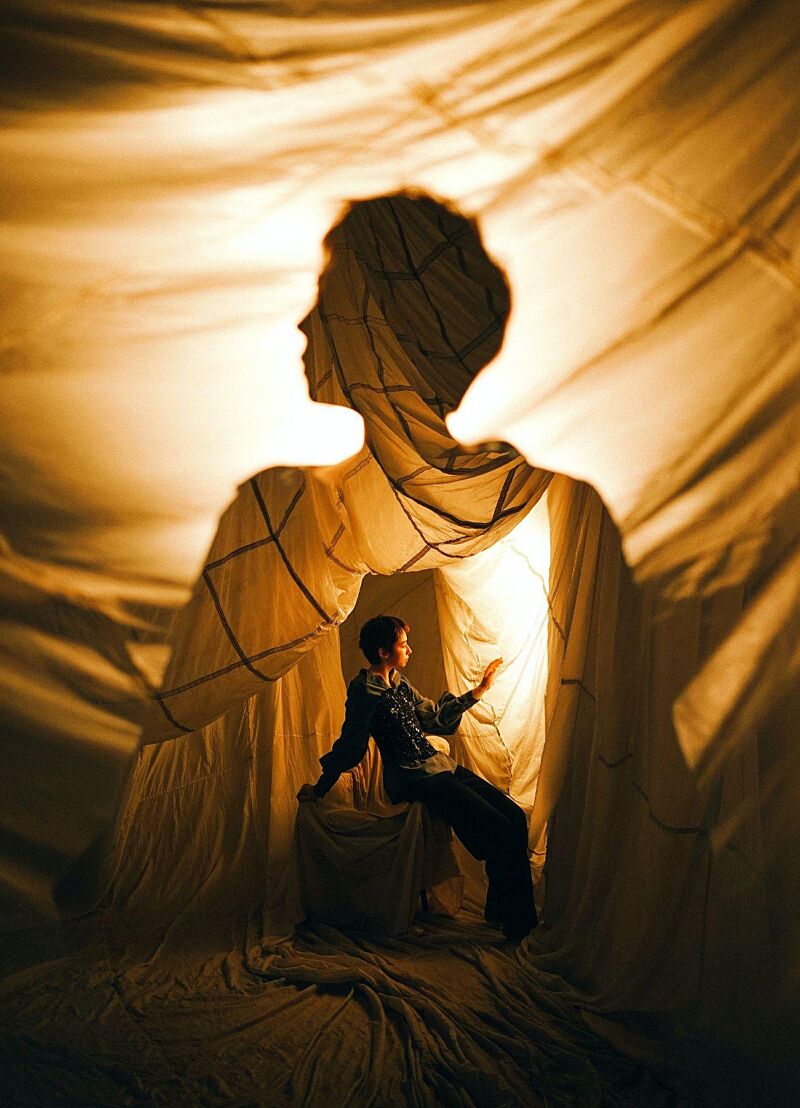 Een jong persoon zit in een tent van lakens. Op de lakens lijkt een digitaal beeld van diezelfde persoon geprojecteerd.