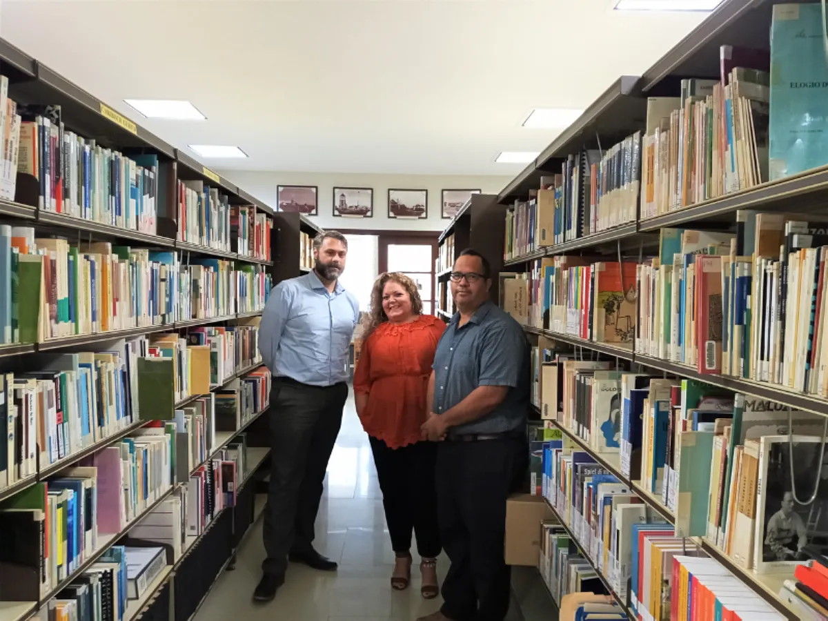 Drie mensen staan in het archief. Tussen twee rijen aan boeken en archiefmateriaal poseren ze voor de camera