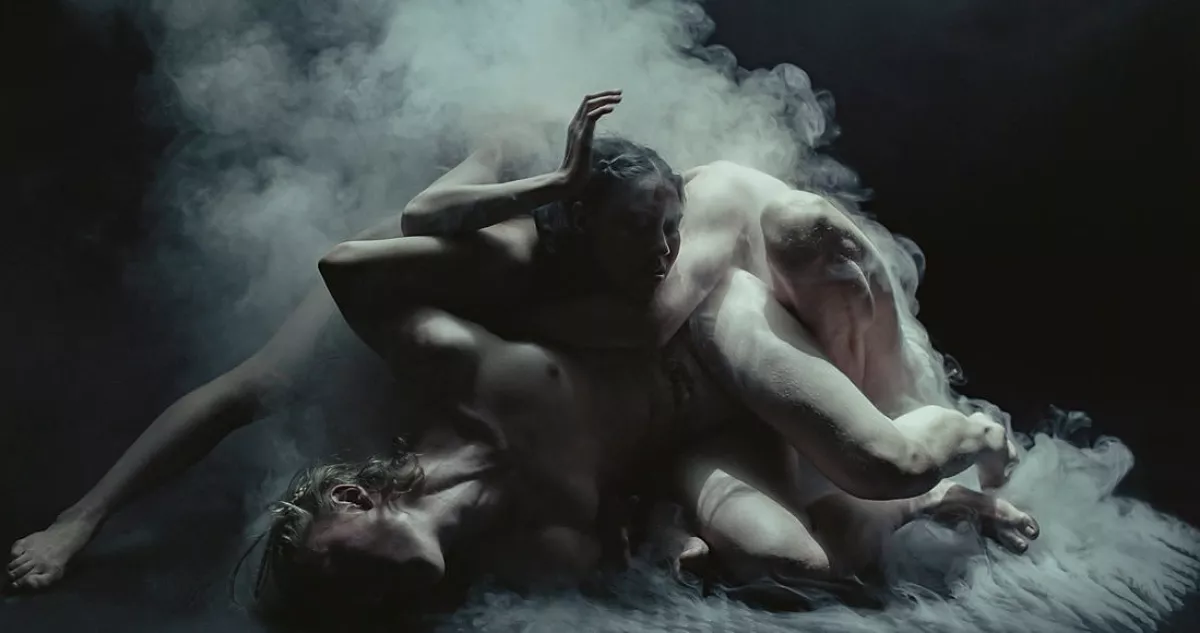 Still from the theaterproduction Mist. Three dancers are moving intertwined with each other across the floor. They're not wearing clothes and are surrounded by smoke or mist.