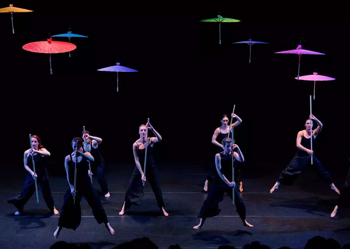 Acht dansers staan in twee rijen op het podium. Ze dragen zwarte kleding en staan tegen een zwarte achtergrond. Boven hen zweven felgekleurde parasols.