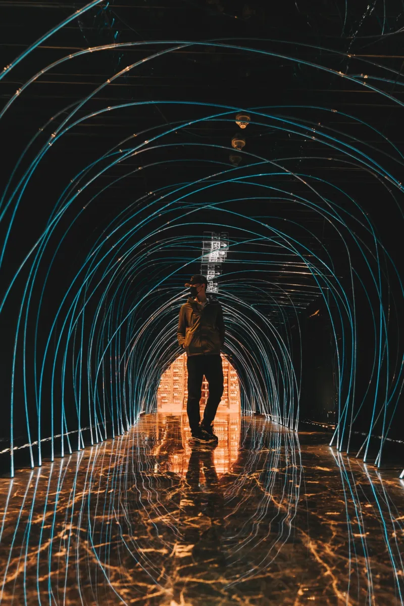 Persoon in een donkere 'tunnel' bestaande uit lichtgevende blauwe bogen. De vloer is zwart met oranje lichtlijnen. Achter in de tunnel staat een soort kast met allemaal kleine lades.