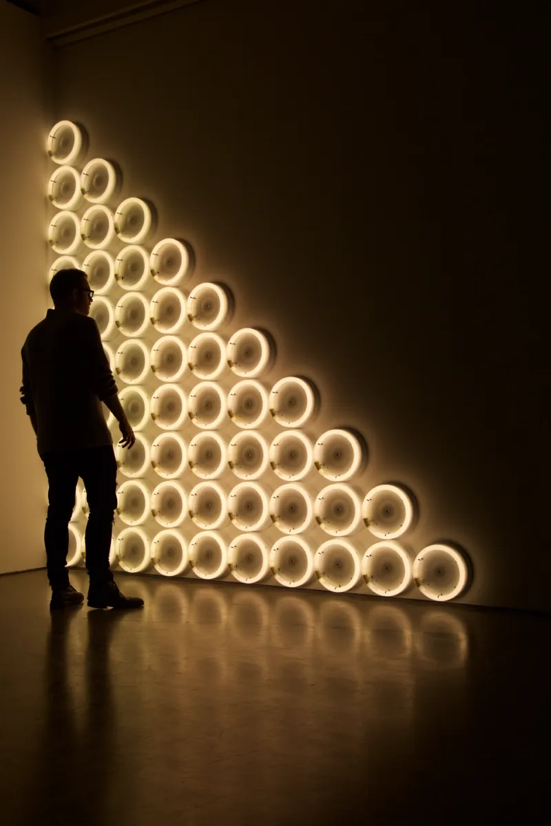 Persoon kijkt naar lichtgevende ringen die aan een muur zijn bevestigd in de vorm van een driehoek. Links tegen de wand zit de hoogste rij en elke rij rechts daarvan heeft één ring minder.