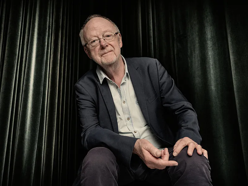 Portret van Louis Andriessen. Een oudere man met bril. Hij draagt een net donkergrijs pak met een wit overhemd. De achtergrond lijkt een donkergrijs theatergordijn. Louis Andriessen lijkt op de rand van een podium te zitten. Het portret toont hem van hoofd tot knieën.