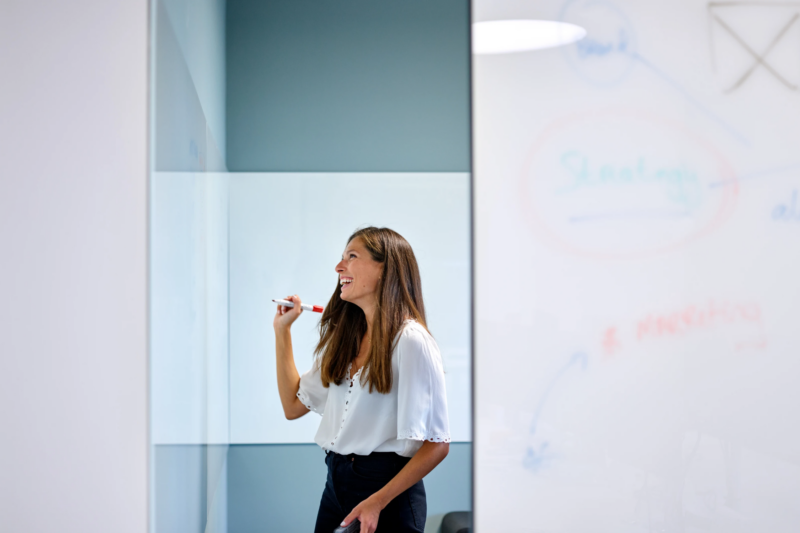 Smiling person with whiteboard marker in hand looks at a whiteboard to brainstorm about digital projects.